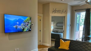 A modern living room with a flat-screen TV, a black sofa with a yellow pillow, a wooden coffee table, and a view of a bedroom with a bed and a 'Home' sign on the wall. - Downtown condo short walk to lake, patio and awesome common area (Grand Lake)