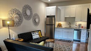 A modern living room with a black sofa, a small round table, and a black lamp. Adjacent to the living room is a kitchen with white cabinets, a stainless steel refrigerator, and a dishwasher. - Downtown condo short walk to lake, patio and awesome common area (Grand Lake)