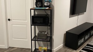 Ein Zimmer mit einer weißen Tür, einer schwarzen Unterhaltungsecke inklusive Flachbildfernseher, einer Kaffeemaschine, einem Mixer, einer Mikrowelle, einem Toaster und einem Mülleimer.