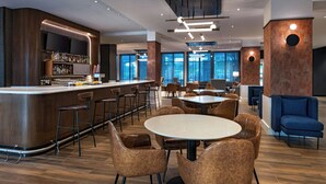 Een moderne bar met een toonbank, barstoelen en een televisiescherm. Er zijn eettafels en stoelen aanwezig, en de ruimte is goed verlicht met plafondlampen.