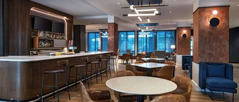 A modern bar area with a counter, bar stools, and a television screen. There are tables and chairs for dining, and the space is well-lit with ceiling lights.
