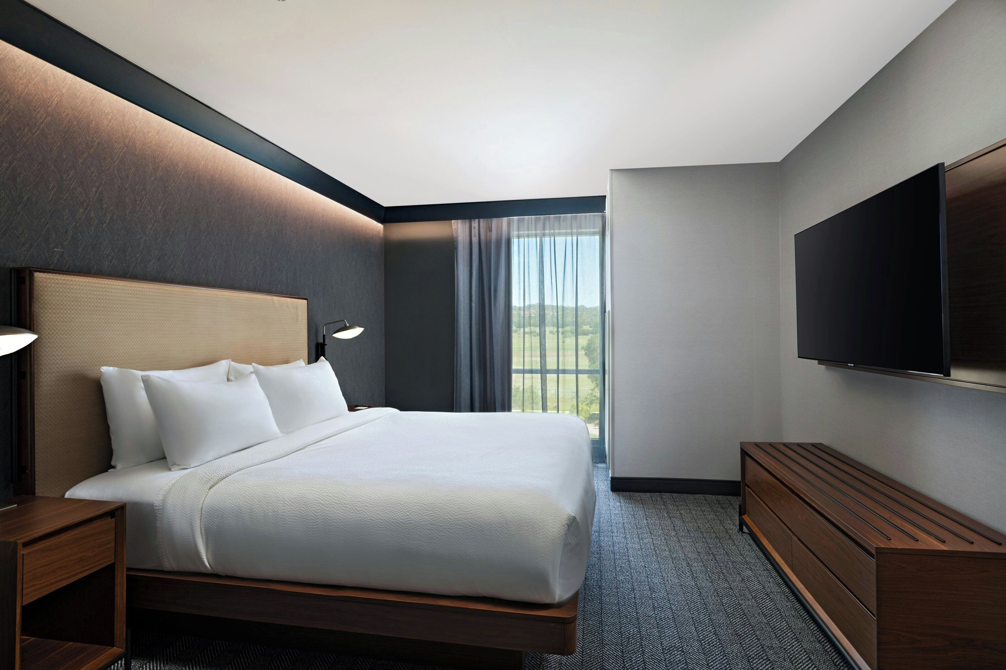 A modern hotel room with a large bed, a TV, and a view of the outdoors.