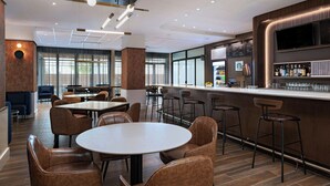 Een modern restaurant met ronde tafels en barstoelen, een toonbank met drankendisplay en grote ramen die voor natuurlijk licht zorgen.