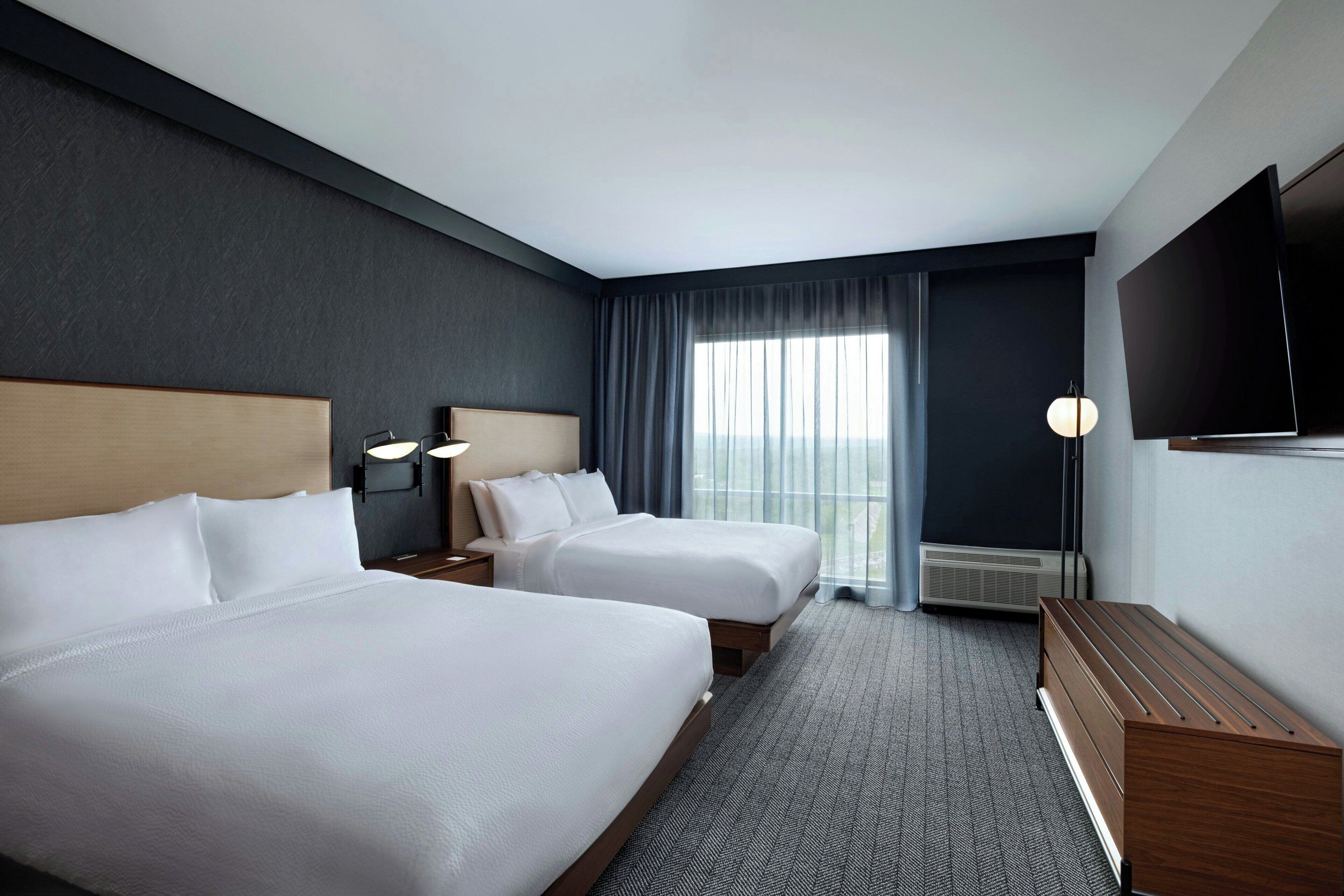 Ett hotellrum med två sängar, en platt-TV, ett skrivbord och ett stort fönster med gardiner.