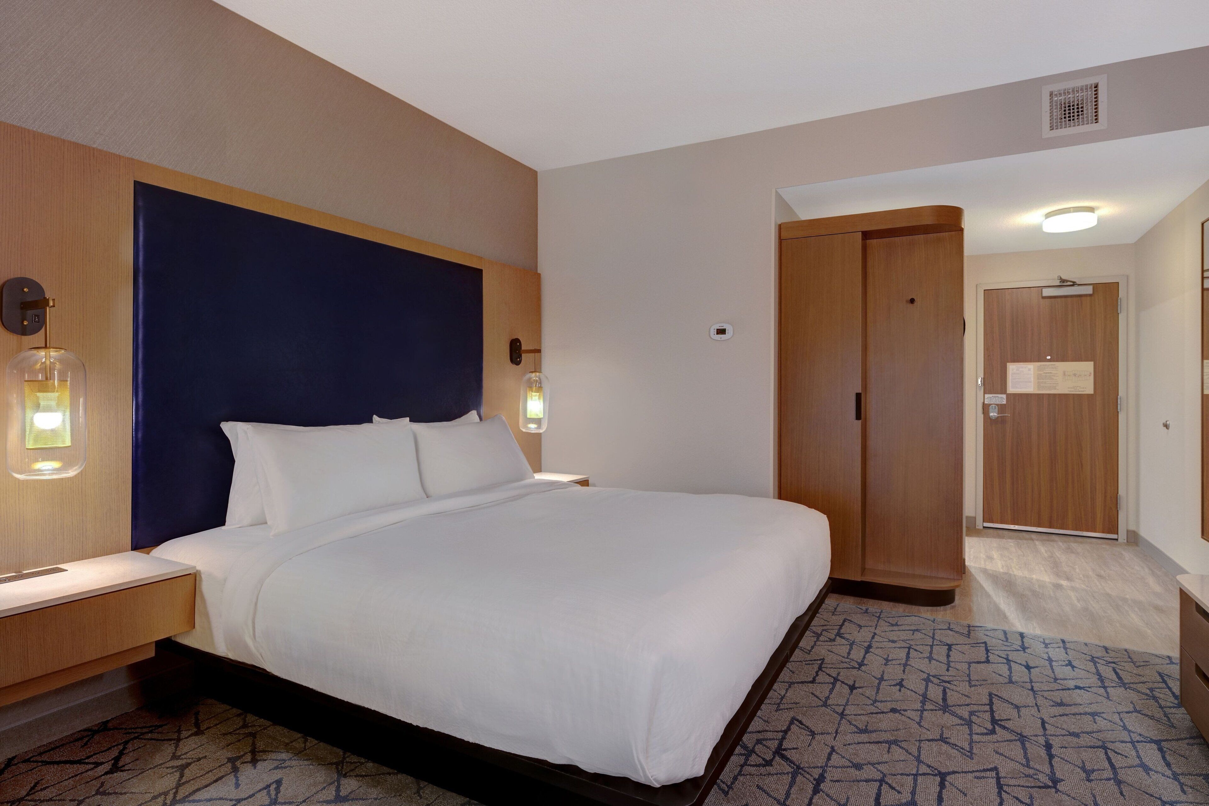 A hotel room with a large bed, wooden headboard, bedside tables with lamps, a patterned rug, and a wardrobe.