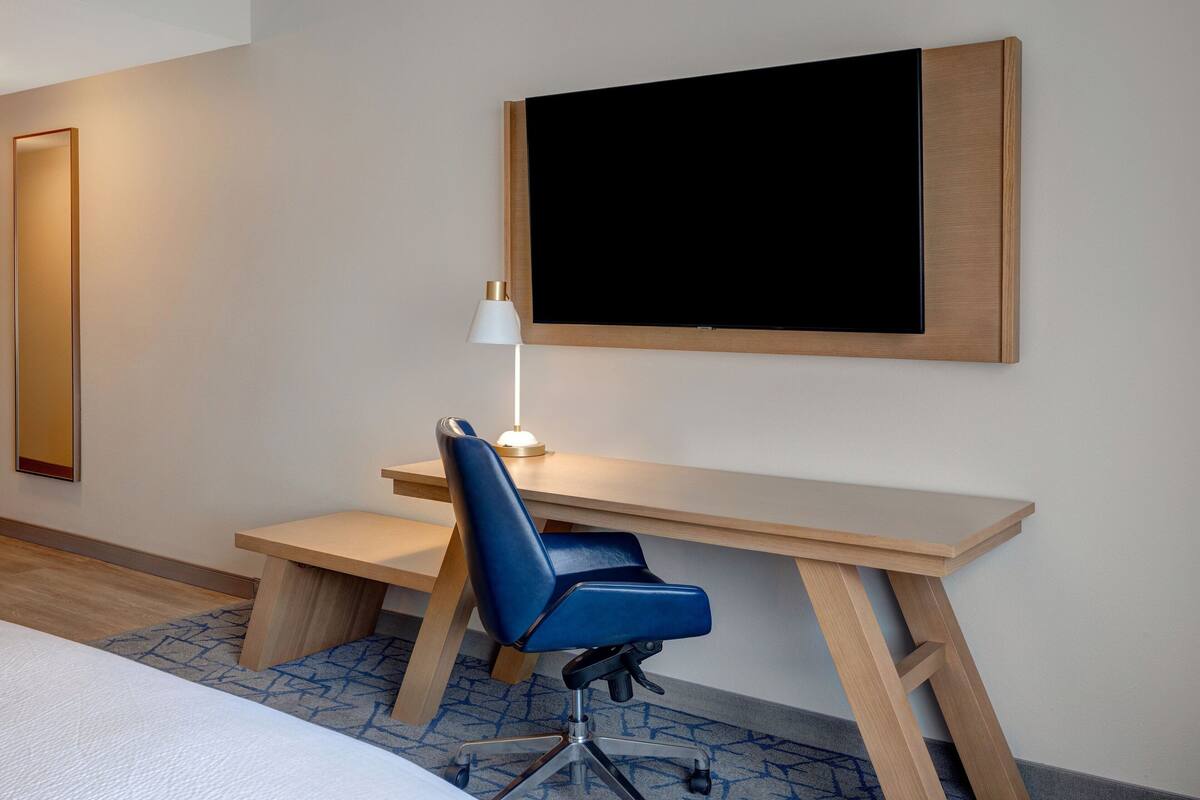 Ein modernes Hotelzimmer mit einem Holztisch, einem blauen Bürostuhl, einer Stehlampe und einem großen, schwarzen Bildschirm an der Wand.