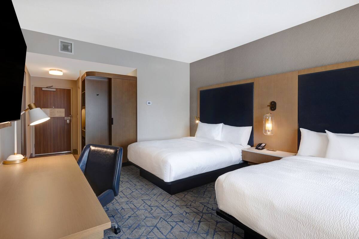 Ein Hotelzimmer mit zwei Betten, einem Schreibtisch mit Lampe, einem Sessel und einer Tür zu einem anderen Raum.