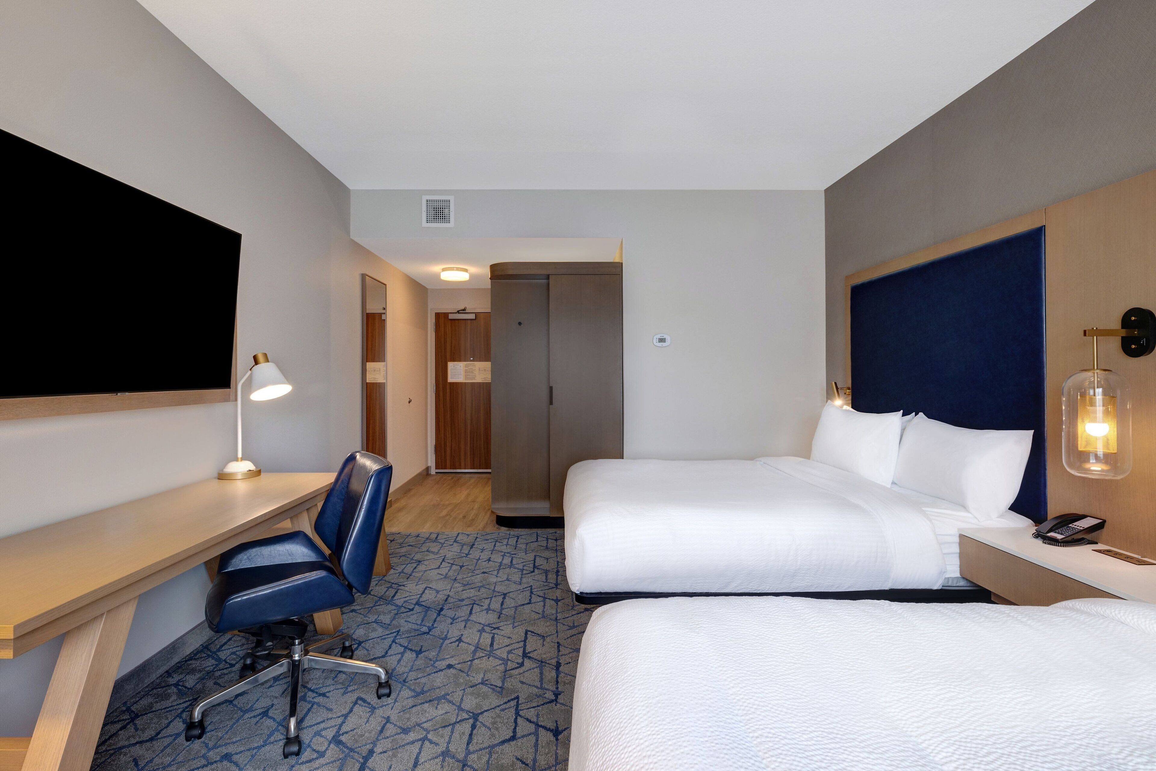 A hotel room with two beds, a desk with a chair, a desk lamp, a telephone, and a wall-mounted light fixture.