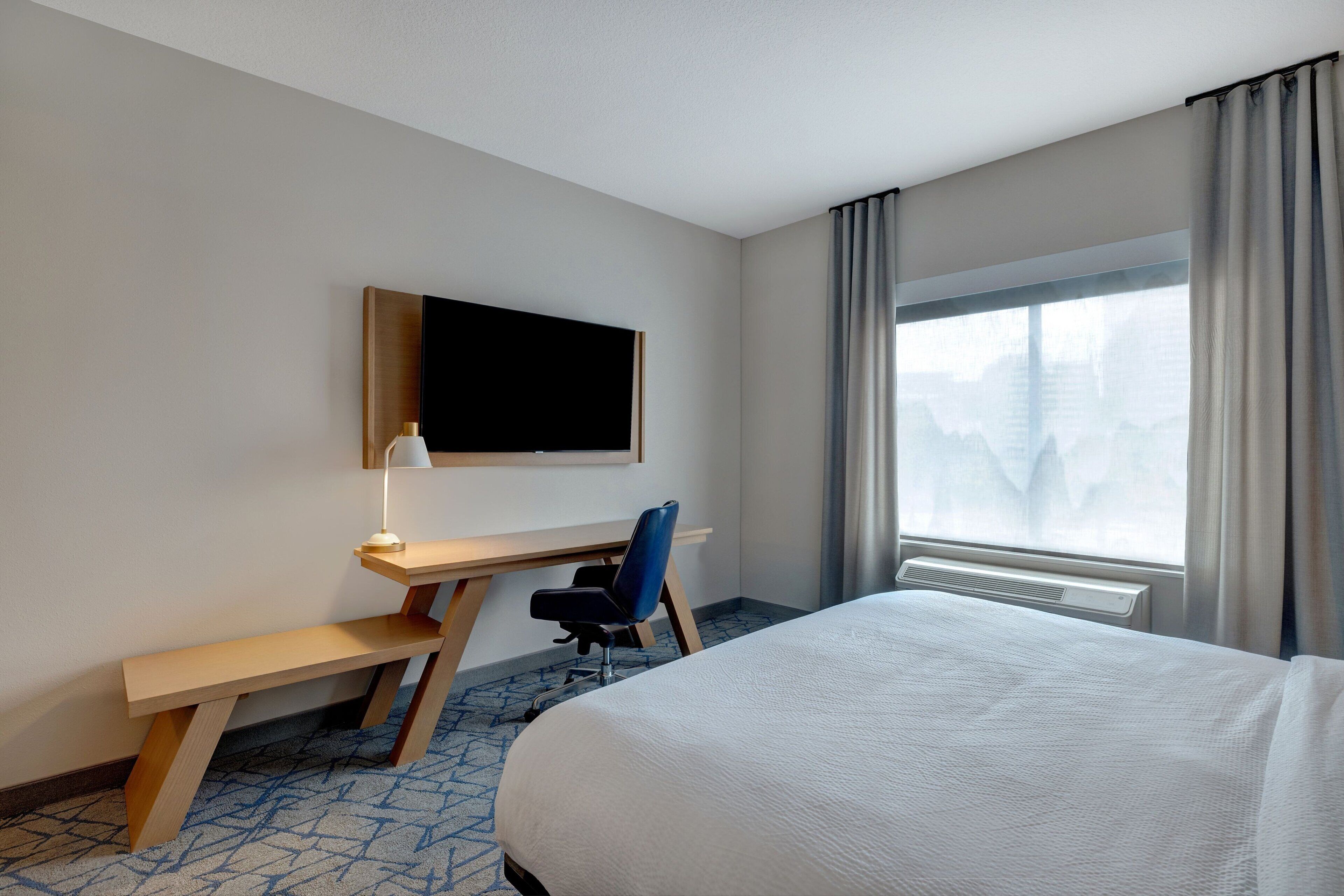 A hotel room with a bed, a desk with a chair, a television, and a window with curtains.