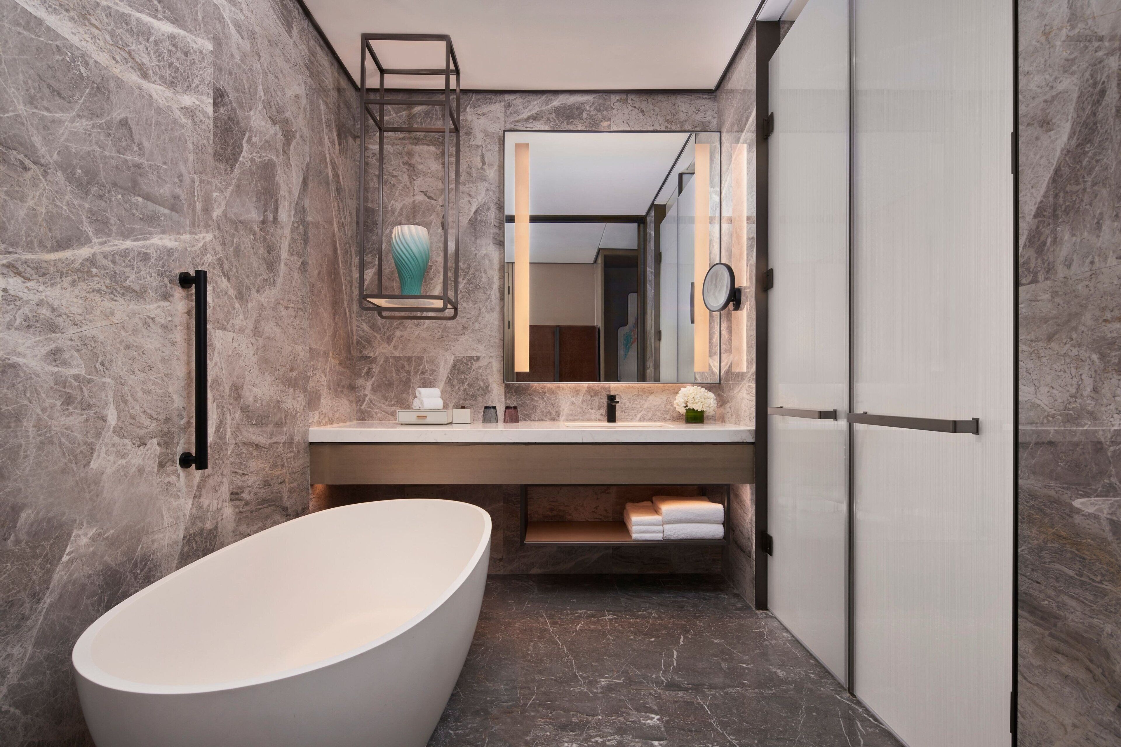 A modern bathroom with a freestanding bathtub, a marble wall, a vanity with a mirror, and a towel rack.