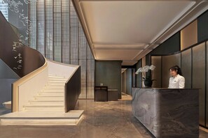 Un moderno vestíbulo de hotel con un mostrador de recepción de mármol, una escalera y una recepcionista.