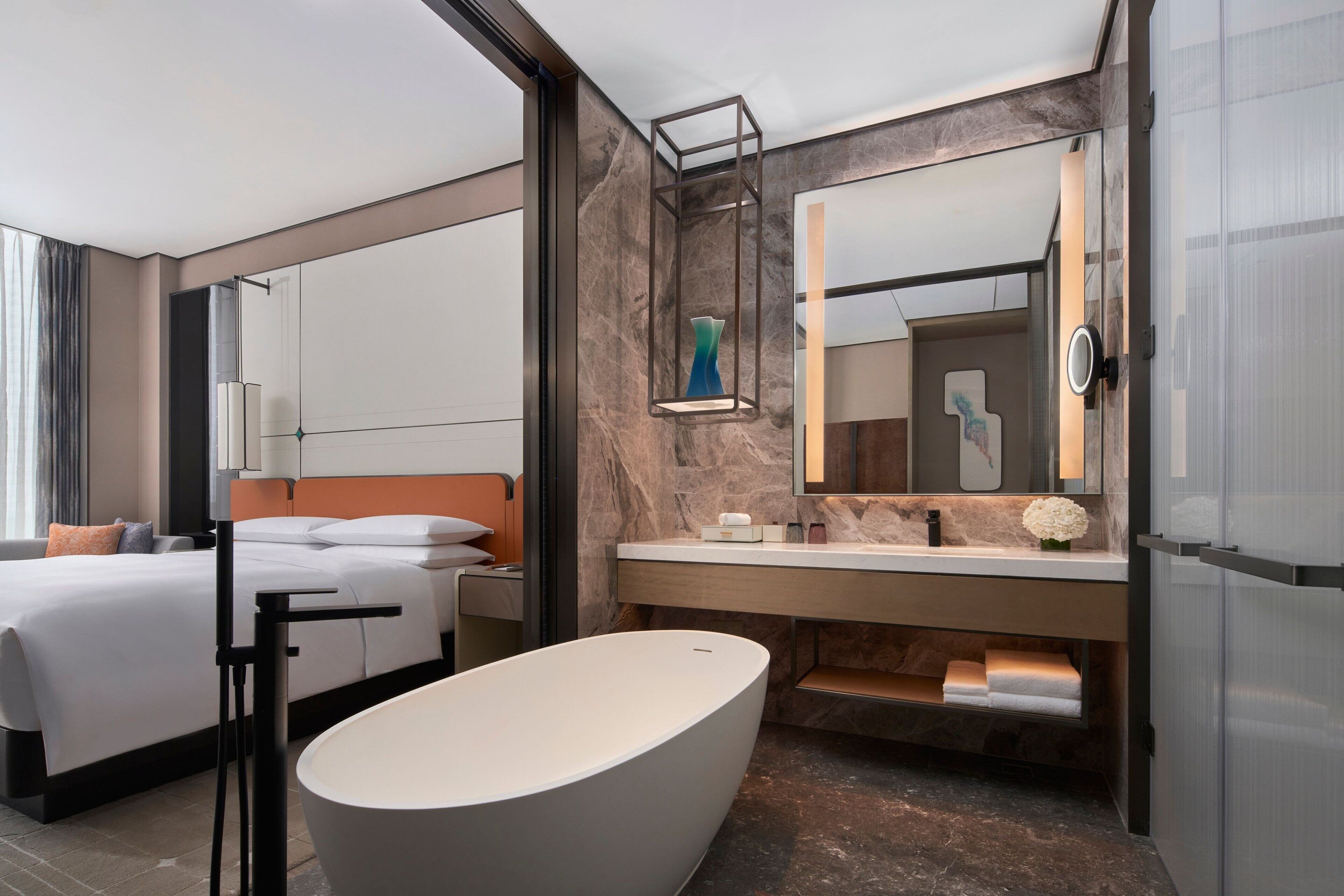 A modern hotel room with a large bed, a freestanding bathtub, a vanity with a mirror, and a large window.