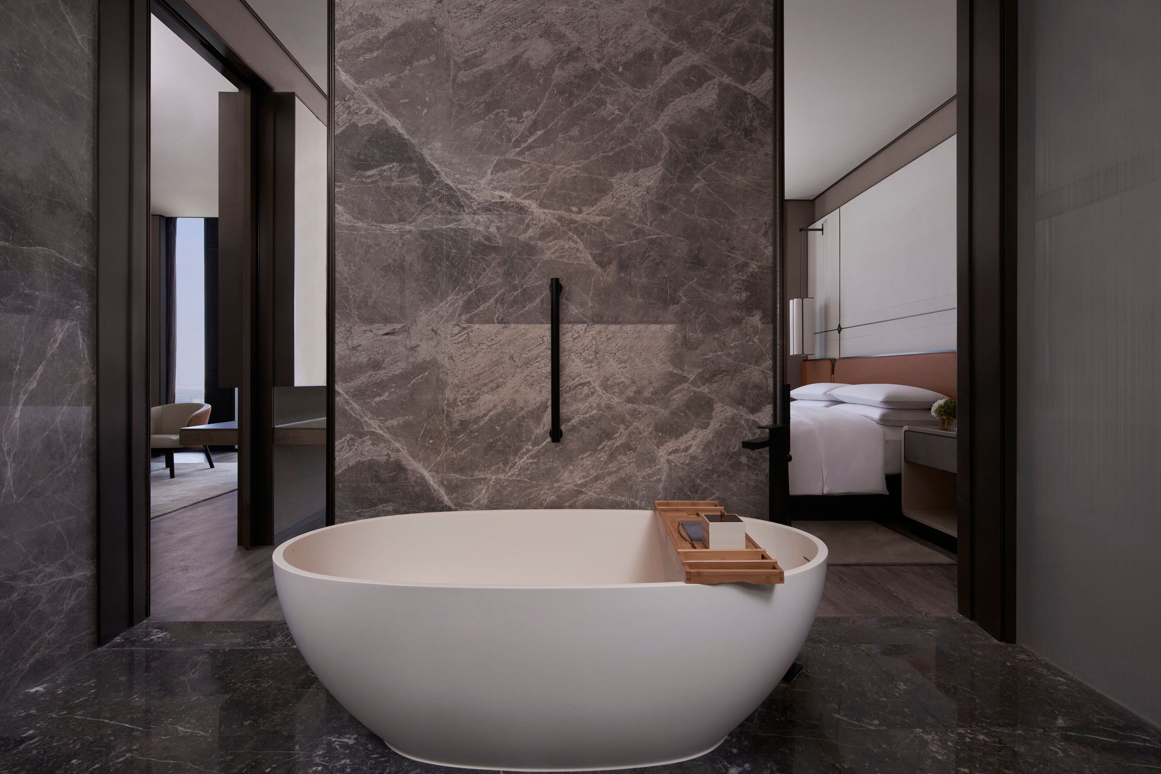 Un baño moderno con una bañera independiente, paredes de mármol y una habitación visible con cama y mesita de noche.