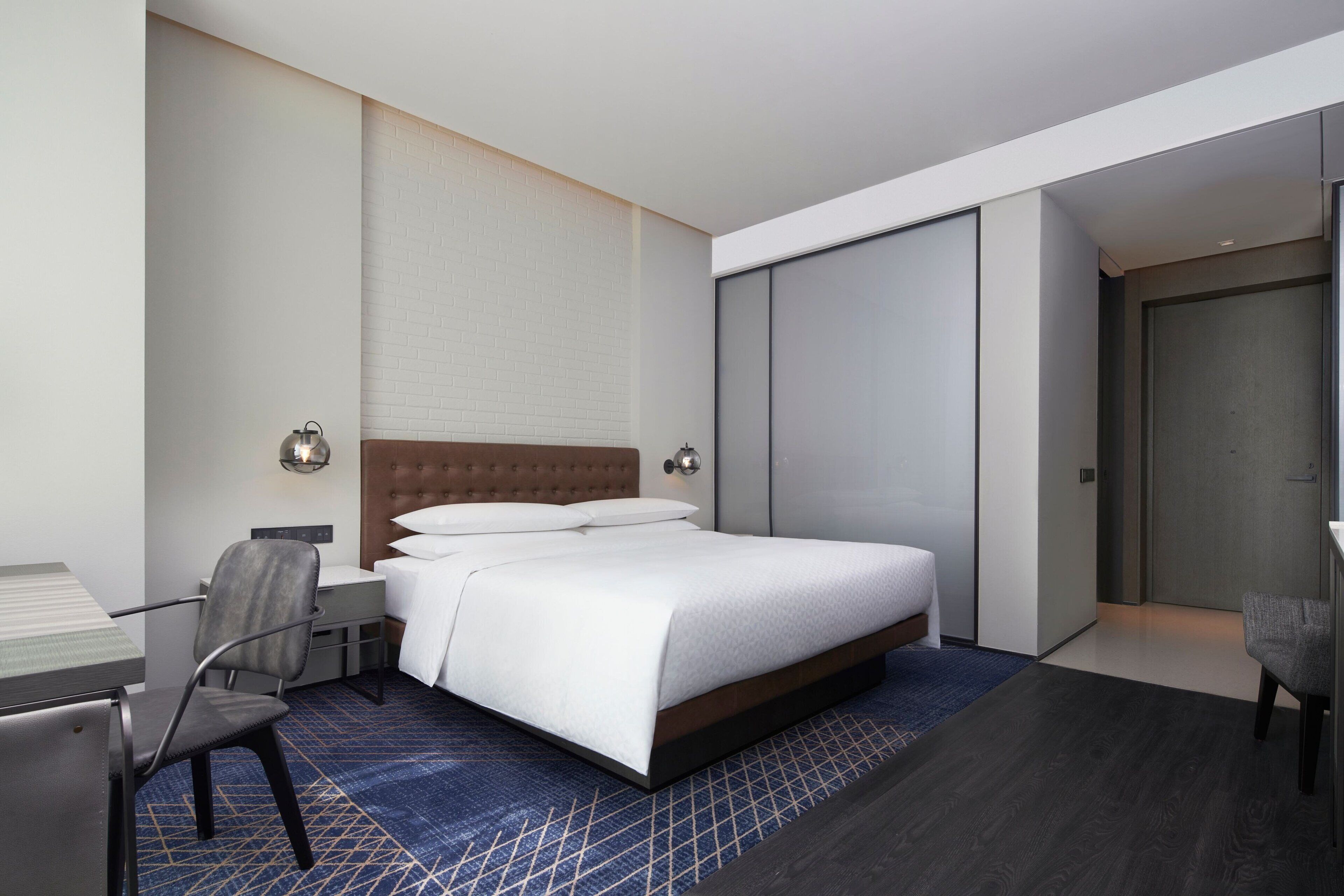 A modern hotel room with a large bed, a desk with a chair, a wardrobe, and a bathroom door.