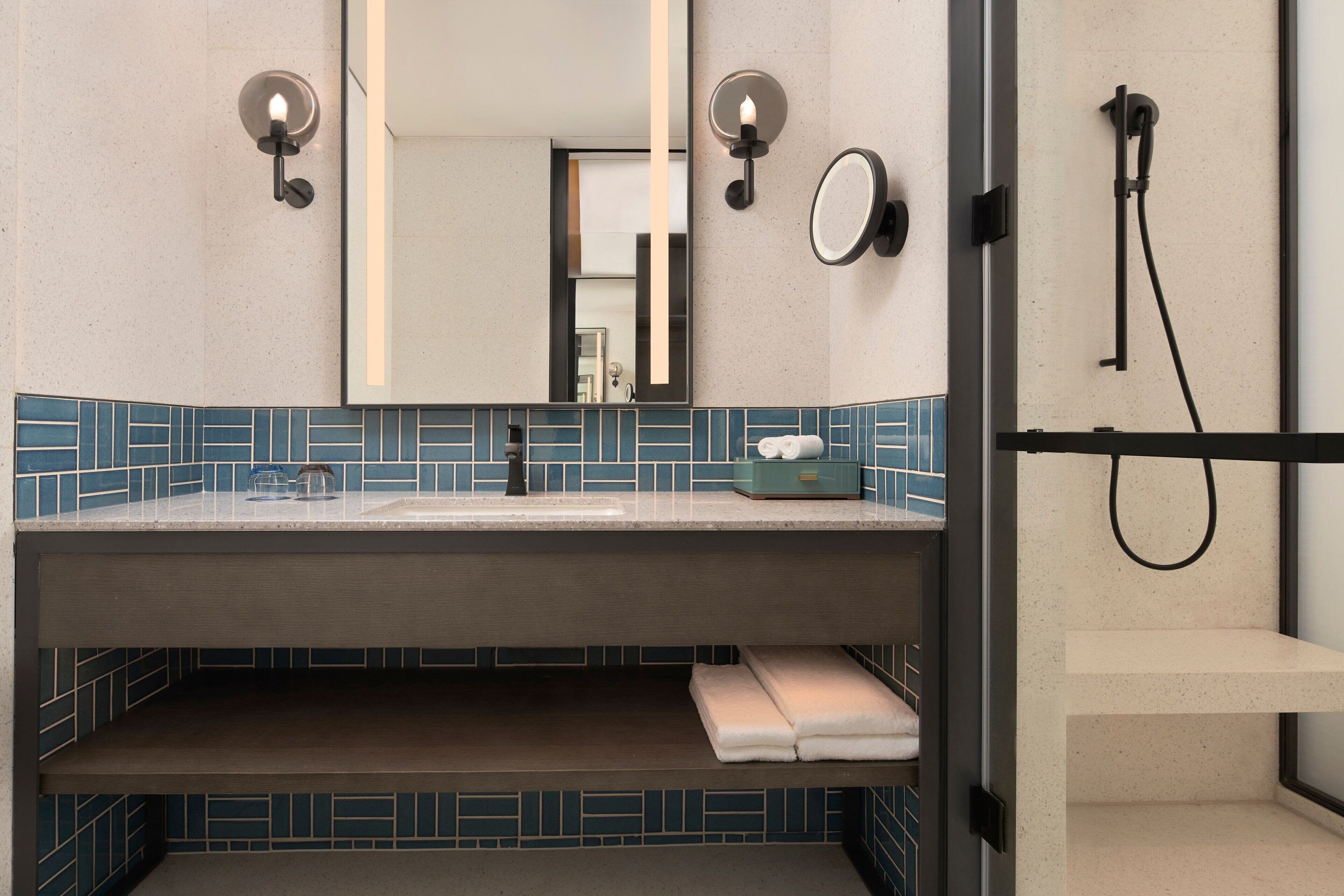 A modern bathroom with a large mirror, two wall-mounted lights, a towel rack, and a shower area.