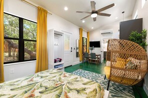 A room with a bed, a ceiling fan, a dining area, and a view of trees.