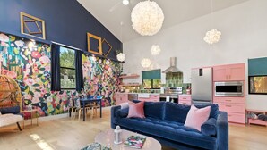 A modern living room with a blue sofa, a dining area with chairs, a kitchen with pink cabinets, and a colorful floral wall. - Treetop House: King Loft & Outdoor Soaking Tub | Wimberley Hill Country (Wimberley)