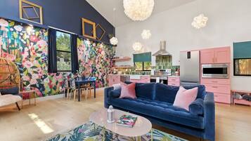 Un salon moderne avec un canapé bleu, un coin repas avec des chaises, une cuisine avec des meubles roses et un mur décoré de motifs floraux colorés.