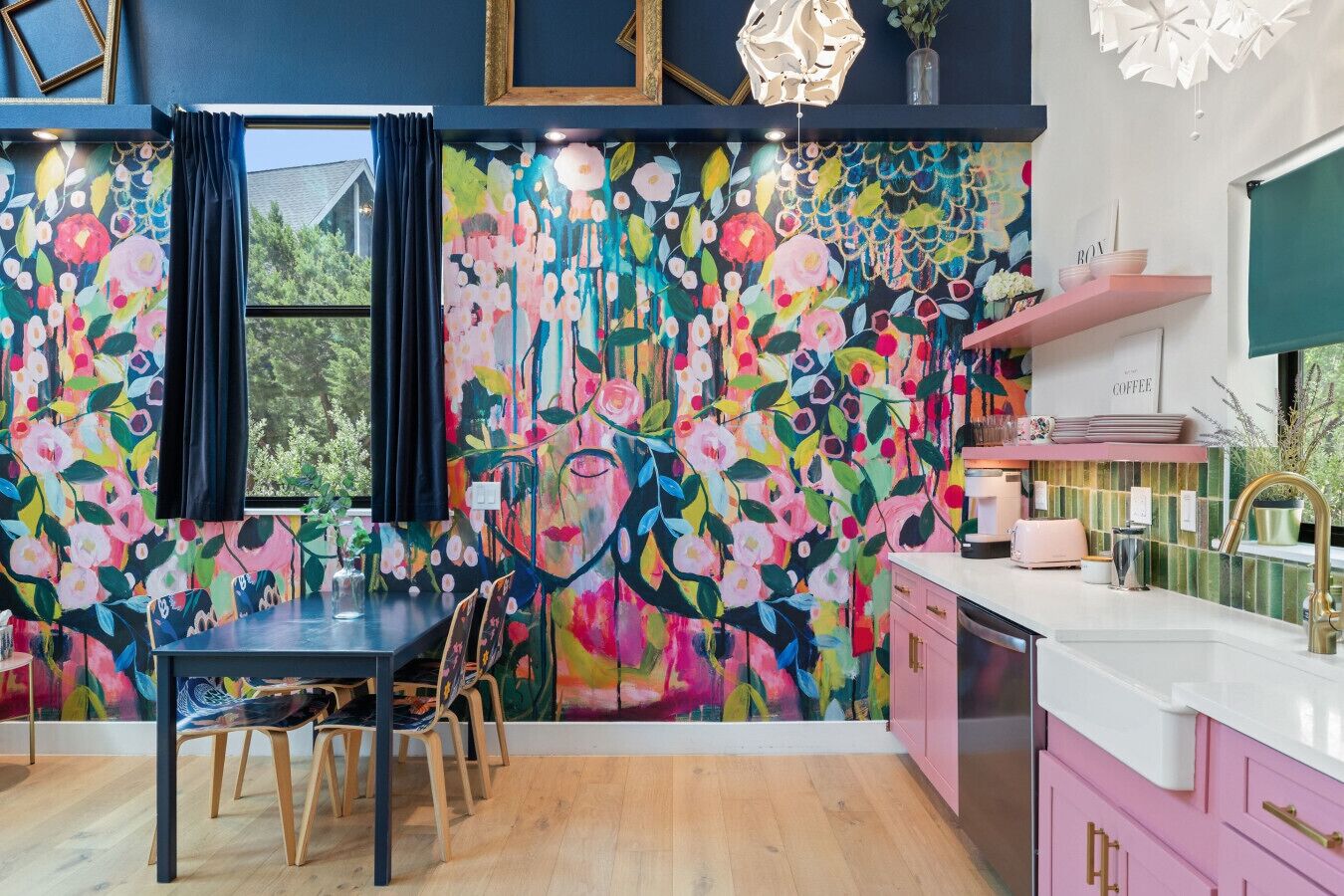 Une cuisine chaleureuse, agrémentée d’une fresque murale aux motifs floraux colorés, d’une table à manger en bois et de placards roses.