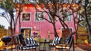 An outdoor seating area with two striped chairs and a small table, set against a pink building with multiple windows. - Treetop House: King Loft & Outdoor Soaking Tub | Wimberley Hill Country (Wimberley)