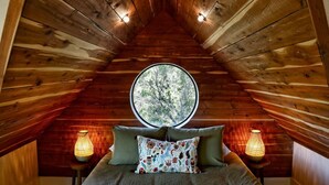 Une chambre confortable située dans les combles, avec un plafond en pente en bois, une fenêtre ronde donnant sur un arbre, un lit avec une literie verte et deux lampes fixées au mur.