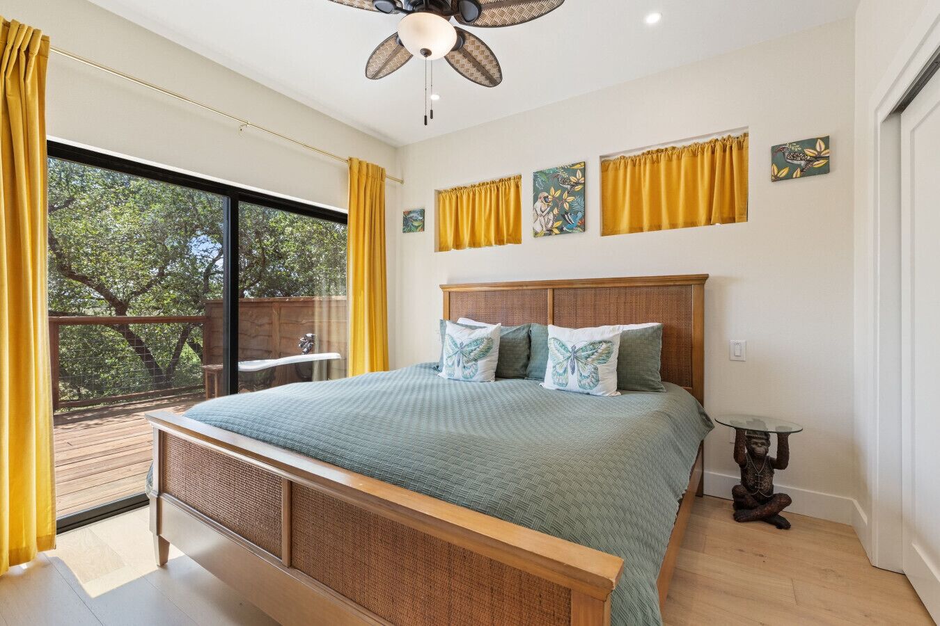 A bedroom with a large bed, a ceiling fan, a view of trees through a sliding door, and a small table with a decorative item.