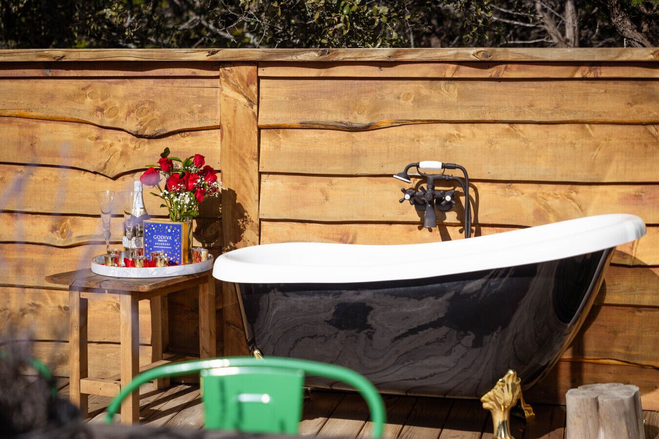 A wooden outdoor hot tub with a black interior, a small table with a bottle and glasses, and a bouquet of red roses.