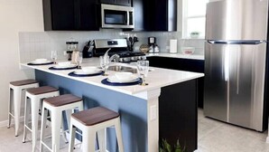 Une cuisine moderne avec des meubles de couleur sombre, un réfrigérateur en acier inoxydable, un four à micro-ondes et une cuisinière à gaz. On y trouve des tabourets de bar et un petit îlot central doté d’un évier.