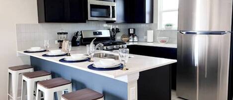 A modern kitchen with dark cabinets, a stainless steel refrigerator, a microwave, and a gas stove. There are bar stools and a small island with a sink.