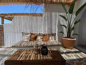 Uma área de estar ao ar livre com iluminação natural, um sofá acolchoado, uma mesa de centro de fibra natural e um vaso com planta.