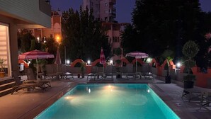 Piscina con sillas de descanso y sombrillas, rodeada de edificios y árboles.