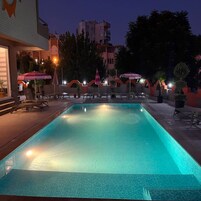 Piscina con sillas de descanso y sombrillas, rodeada de edificios y árboles.