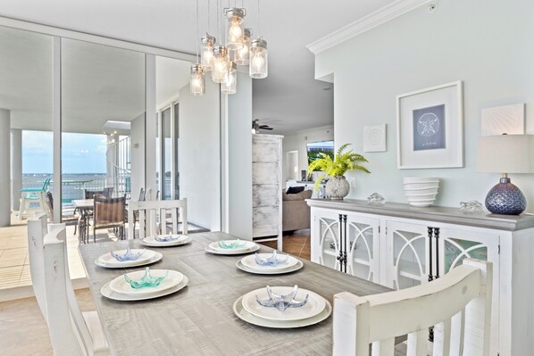 A modern dining area with a long table, white chairs, and a view of the ocean.