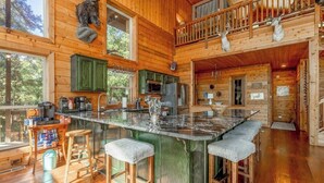 Una cabaña espaciosa con interior de madera, una isla central con taburetes de bar, zona de cocina y un balcón con barandillas de madera.