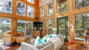 Una cabaña de madera espaciosa con amplias ventanas, un espacio de sala acogedor con un sofá y sillones, y una zona de cocina visible.