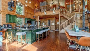 Interior espacioso de una cabaña con una barra central tipo isla, pisos de madera y una escalera que sube a un nivel superior.