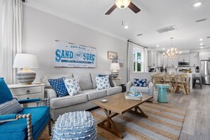 Un salón moderno con un sofá gris, un sillón azul y una mesa de centro de madera. Se ve una cocina al fondo con armarios blancos y electrodomésticos de acero inoxidable.