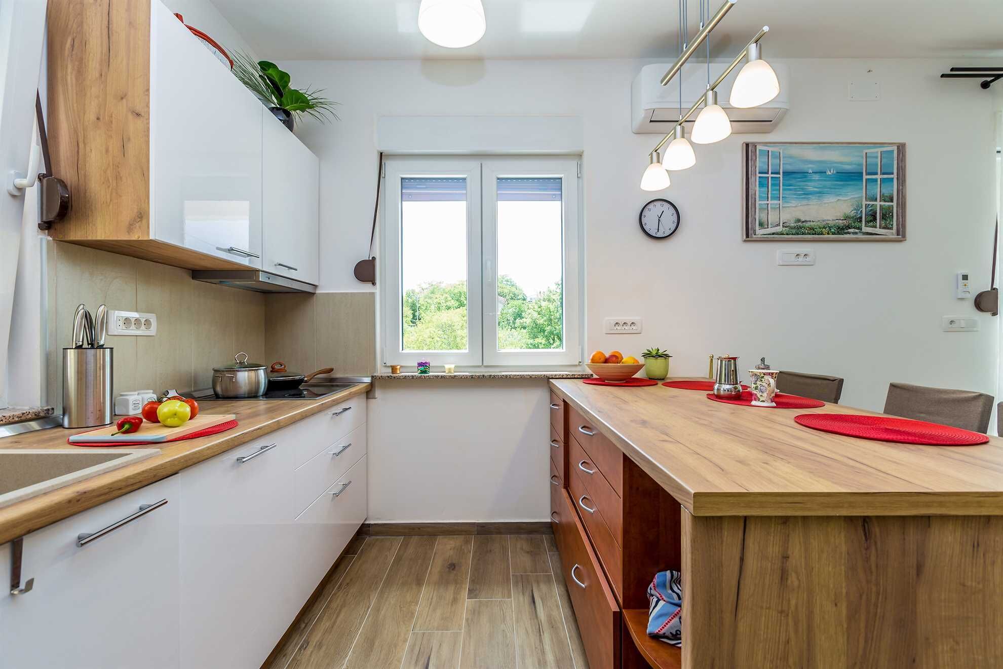 A modern kitchen with white cabinets, a wooden countertop, and a window with a view of greenery. There is a clock on the wall and a painting above the window.