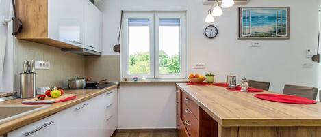 A modern kitchen with white cabinets, a wooden countertop, and a window with a view of greenery. There is a clock on the wall and a painting above the window.