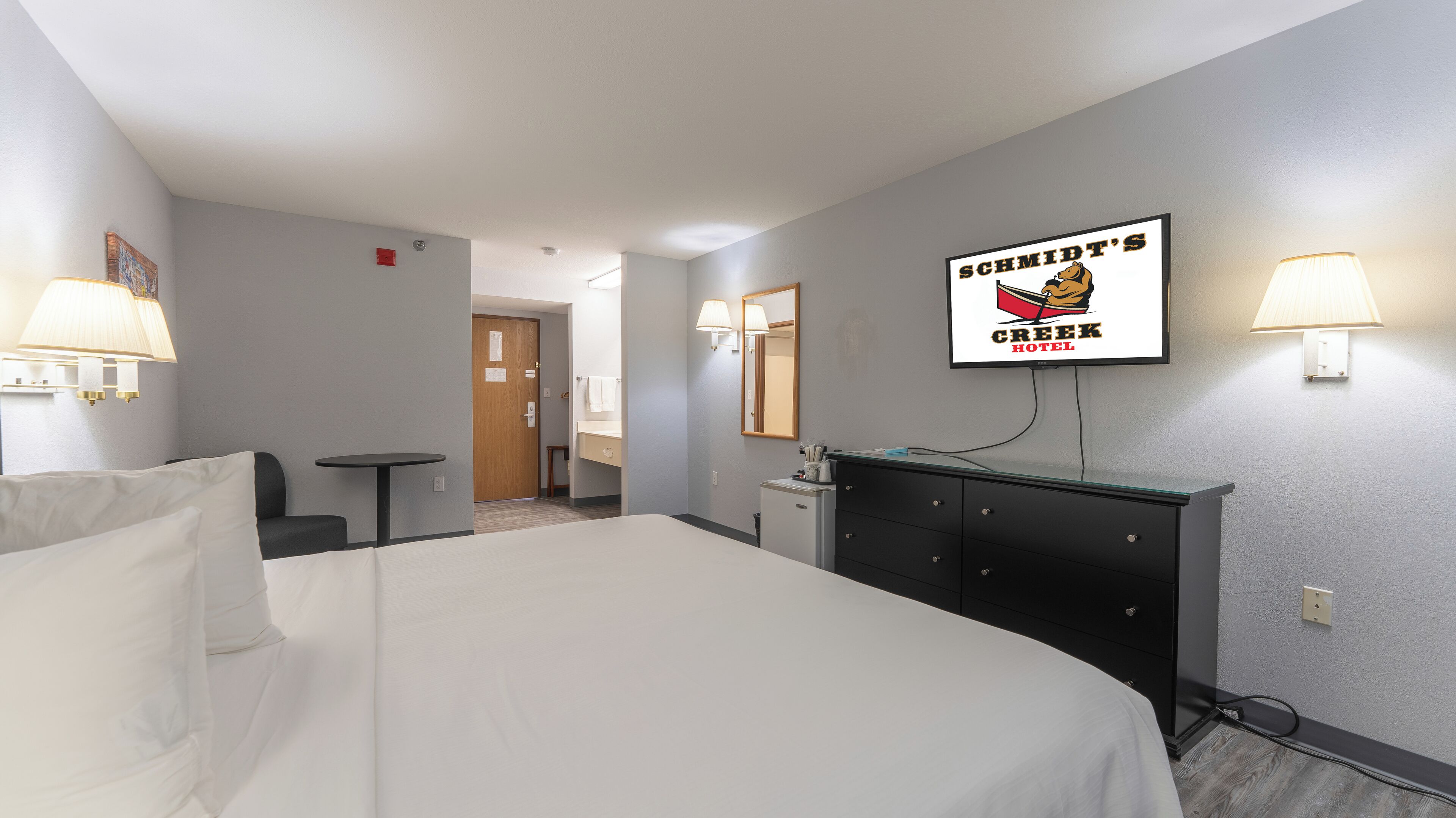 A hotel room with a bed, a desk, a chair, a dresser, and a television displaying the hotel's name.