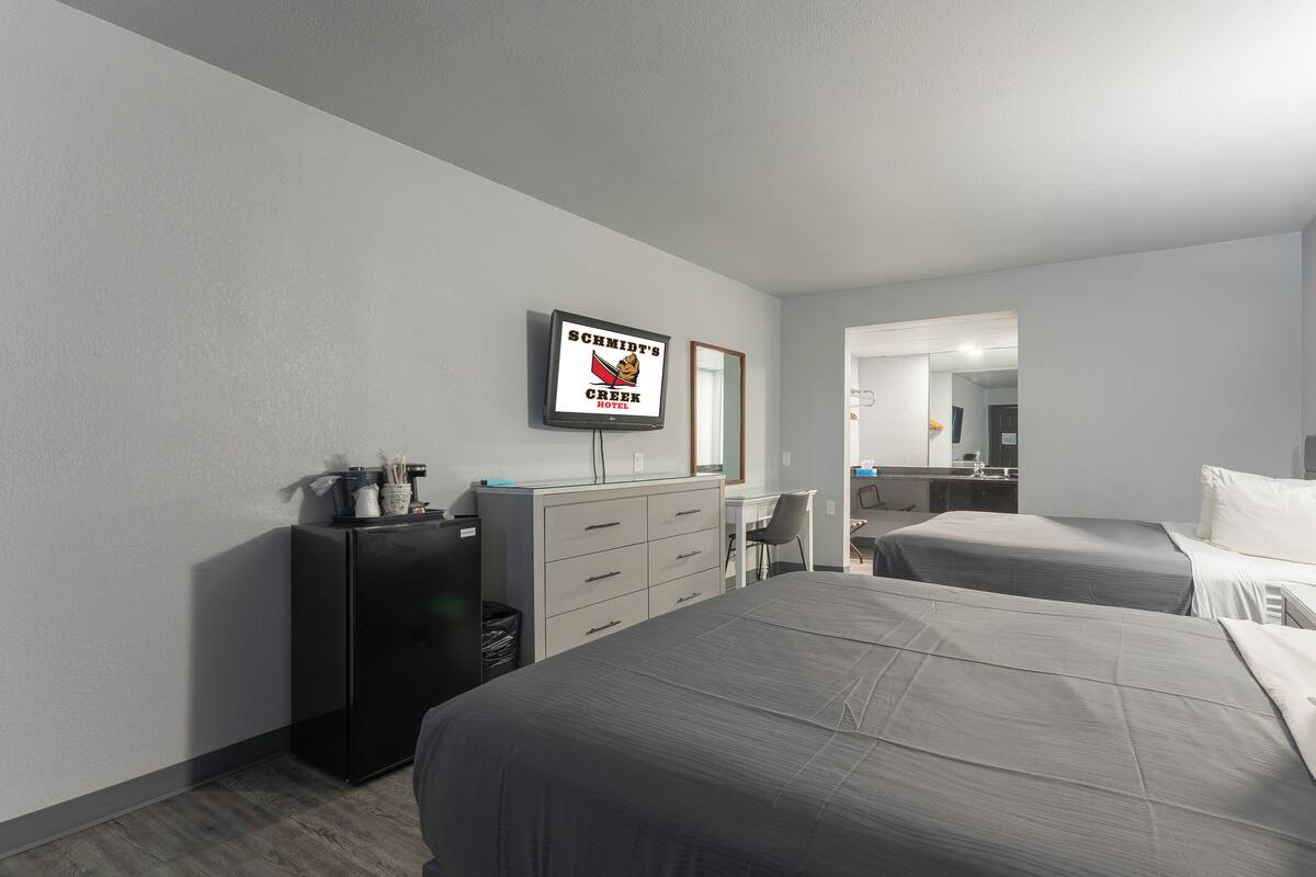 Une chambre d’hôtel avec deux lits, un bureau, un réfrigérateur, un miroir et un téléviseur diffusant l’image de l’enseigne « SCHMIDT’S CREPE HOTEL ».