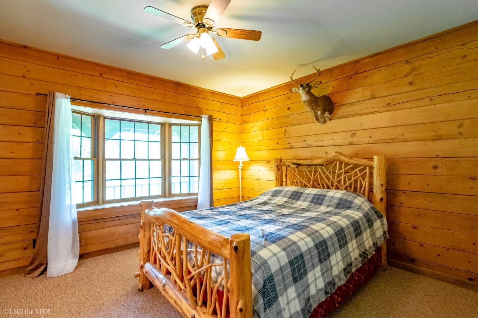 A wooden cabin bedroom with a bed, a ceiling fan, a window, and a mounted deer head.