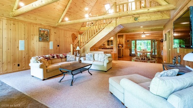 A spacious log cabin interior with a high ceiling, a staircase leading to an upper level, and a cozy living area with sofas and a coffee table.