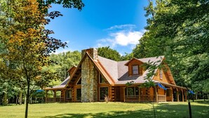 A log cabin with a stone chimney, surrounded by a grassy yard and trees.