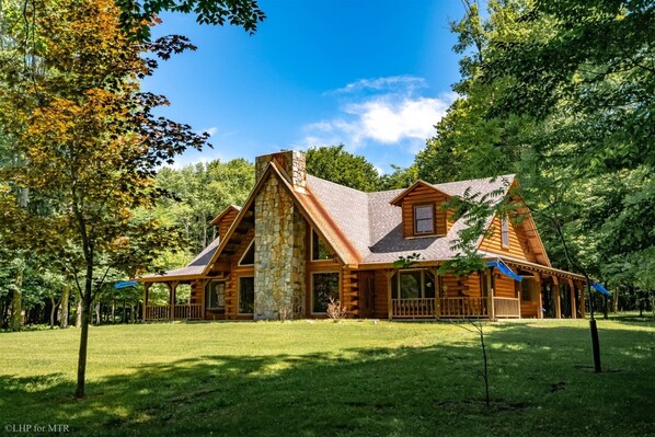 A log cabin with a stone chimney, surrounded by a grassy yard and trees.