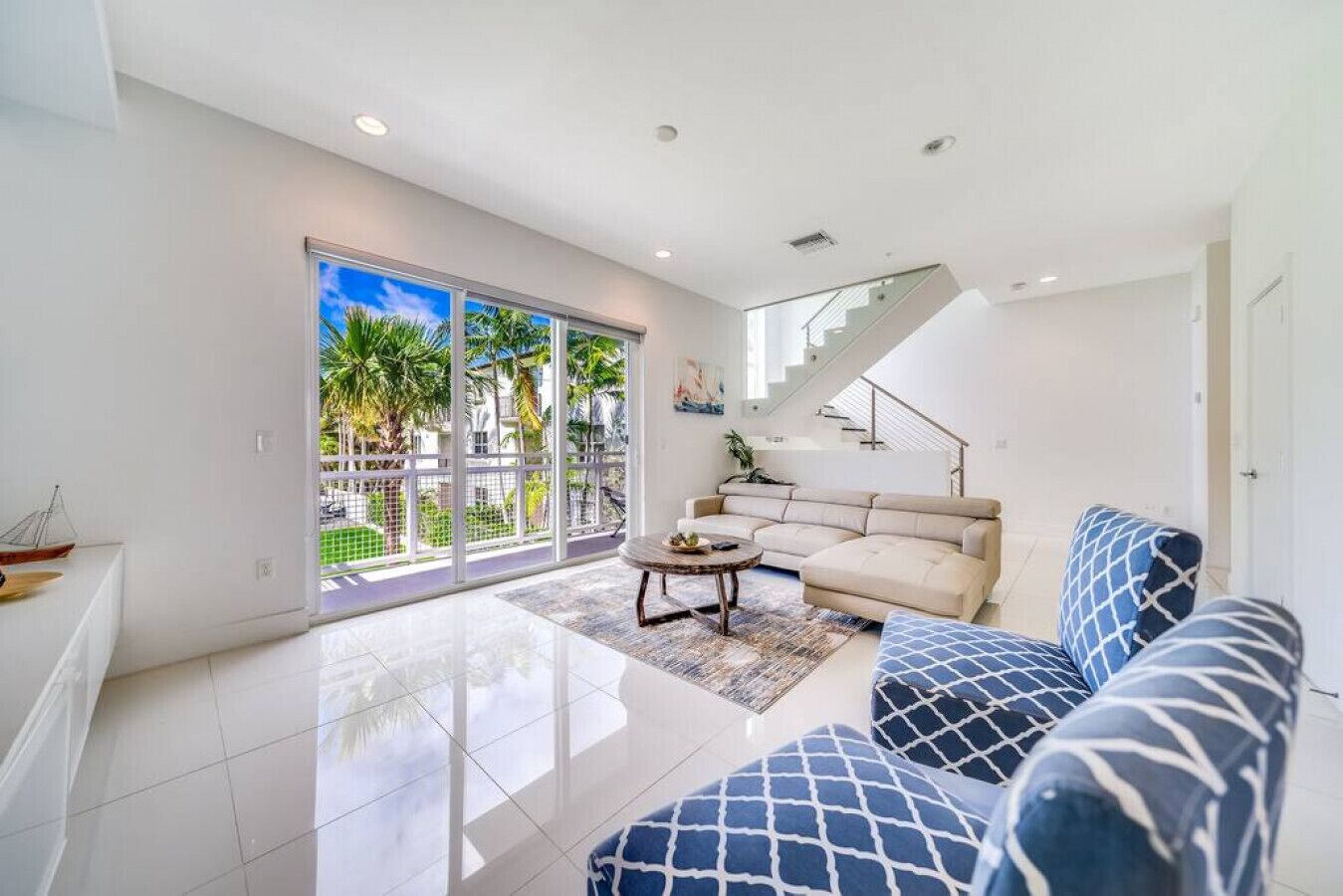 A spacious living area with a large sliding glass door leading to a balcony, a beige sectional sofa, a patterned armchair, and a staircase.