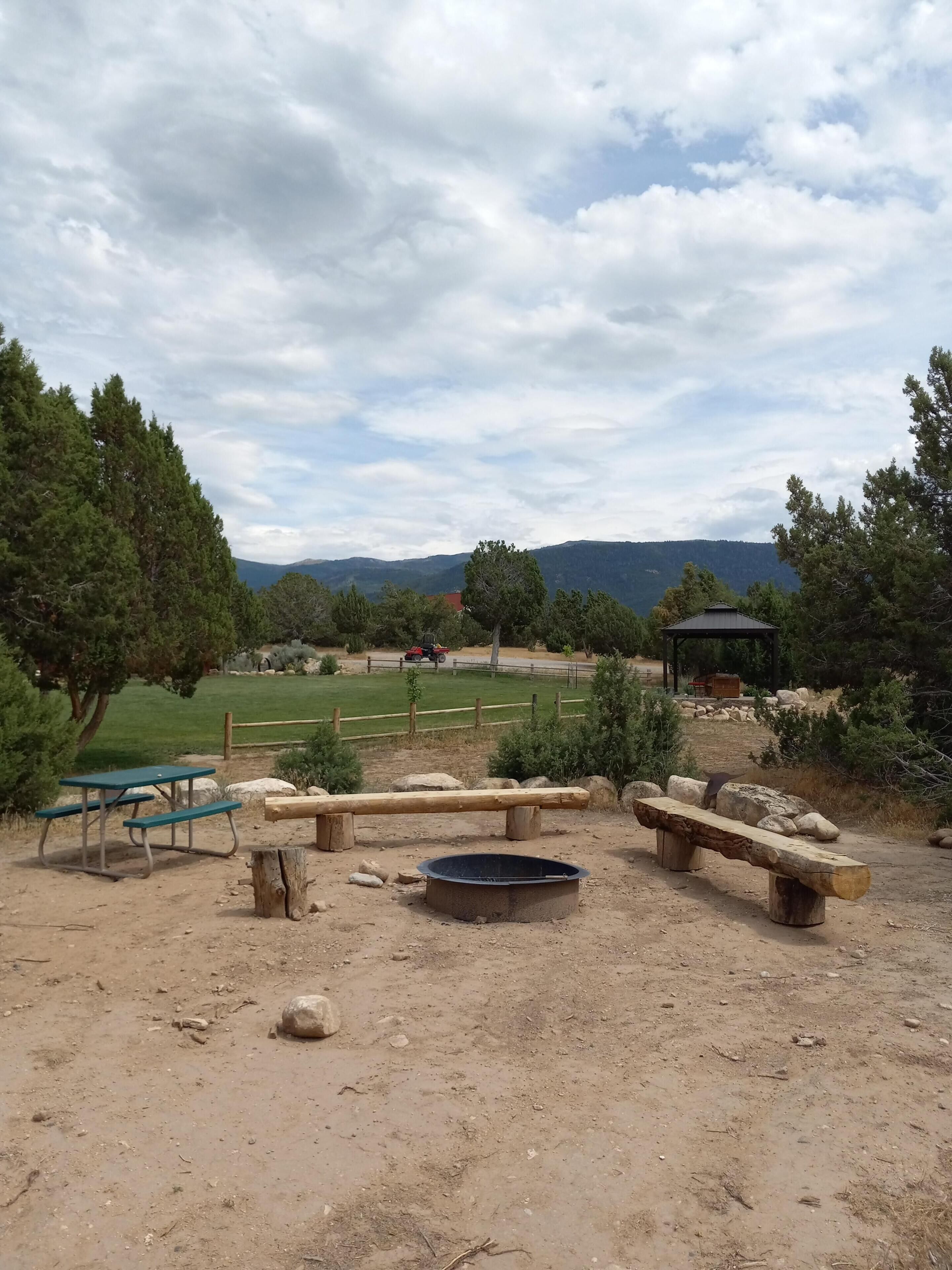 A picnic area with benches and a table, surrounded by trees and a view of distant hills.