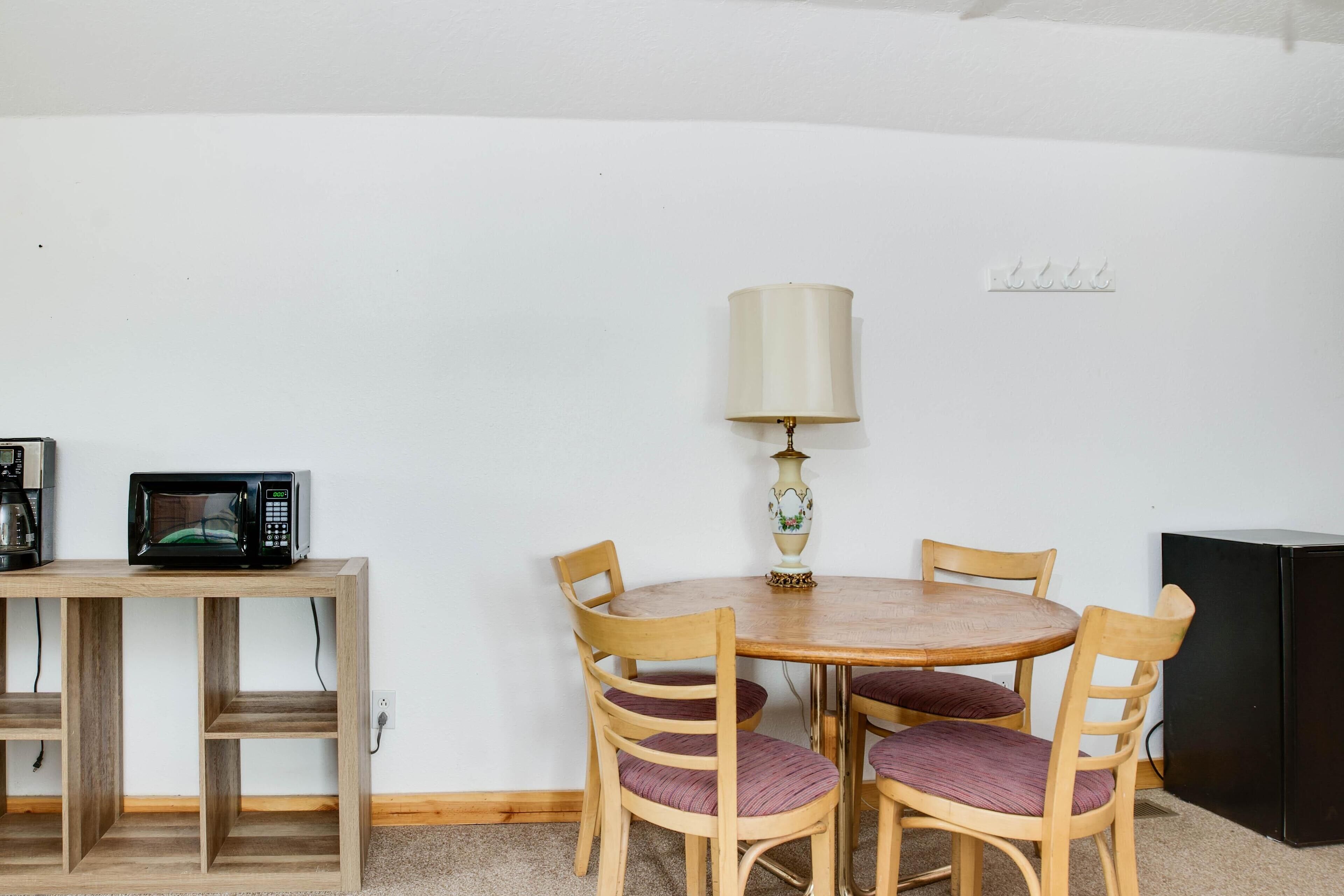 Un comedor con una mesa redonda de madera, cuatro sillas de madera con cojines morados, una lámpara, un microondas y un refrigerador negro.