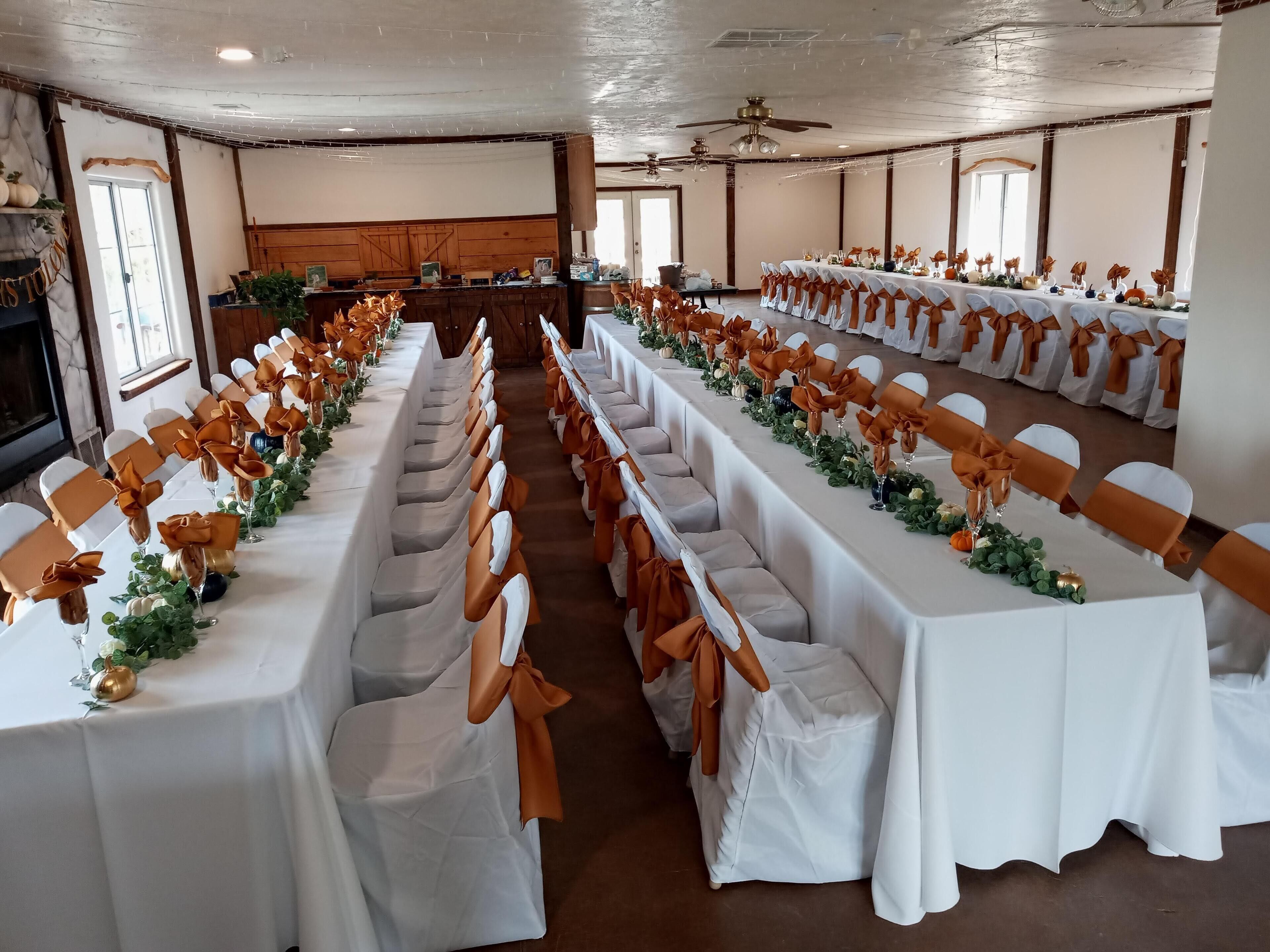 Une salle de banquet avec de longues tables, des nappes blanches et des rubans orange sur les chaises.