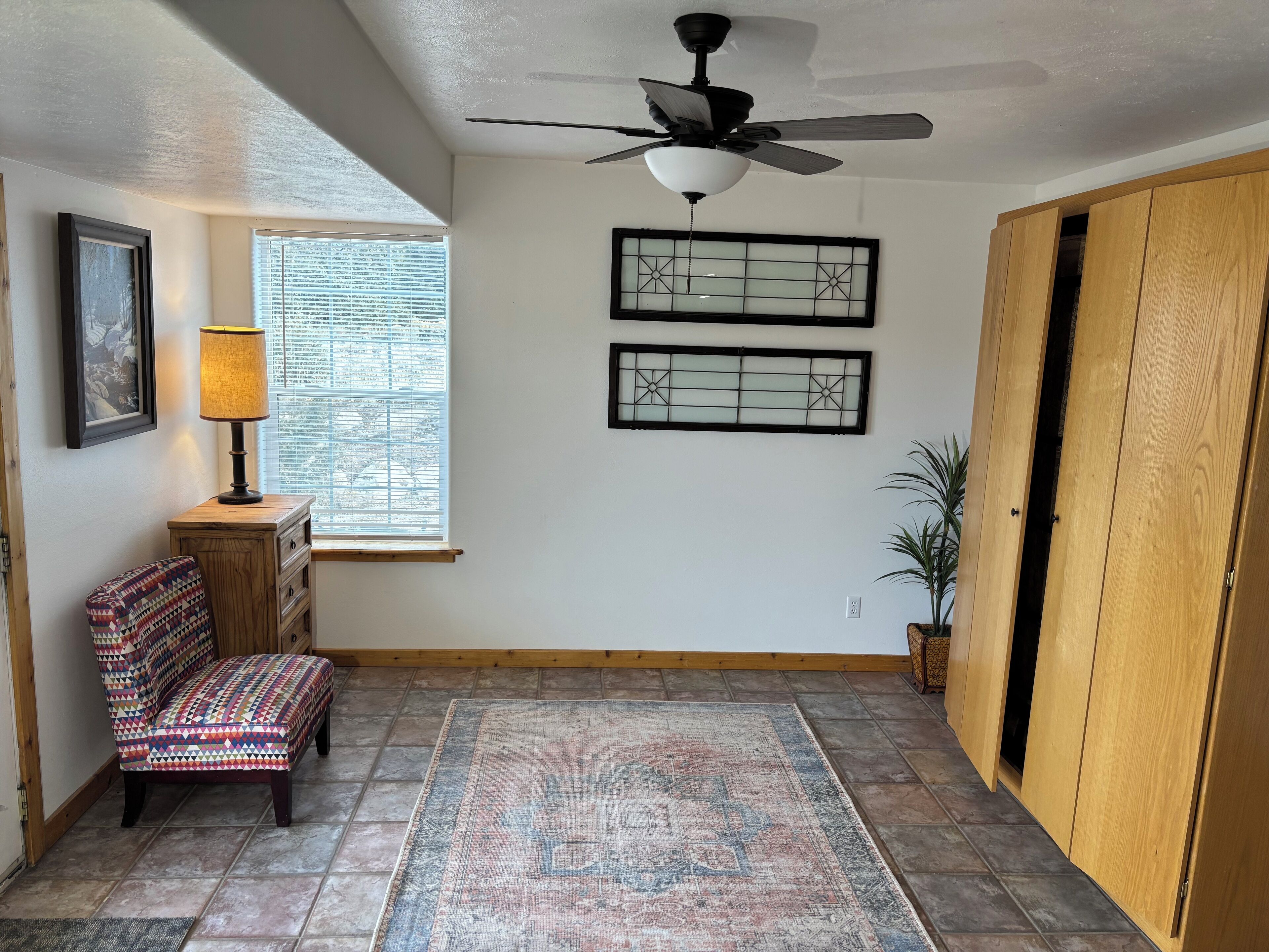 Une pièce avec un ventilateur de plafond, un tapis à motifs, un banc, une fenêtre avec des stores et une porte en bois.